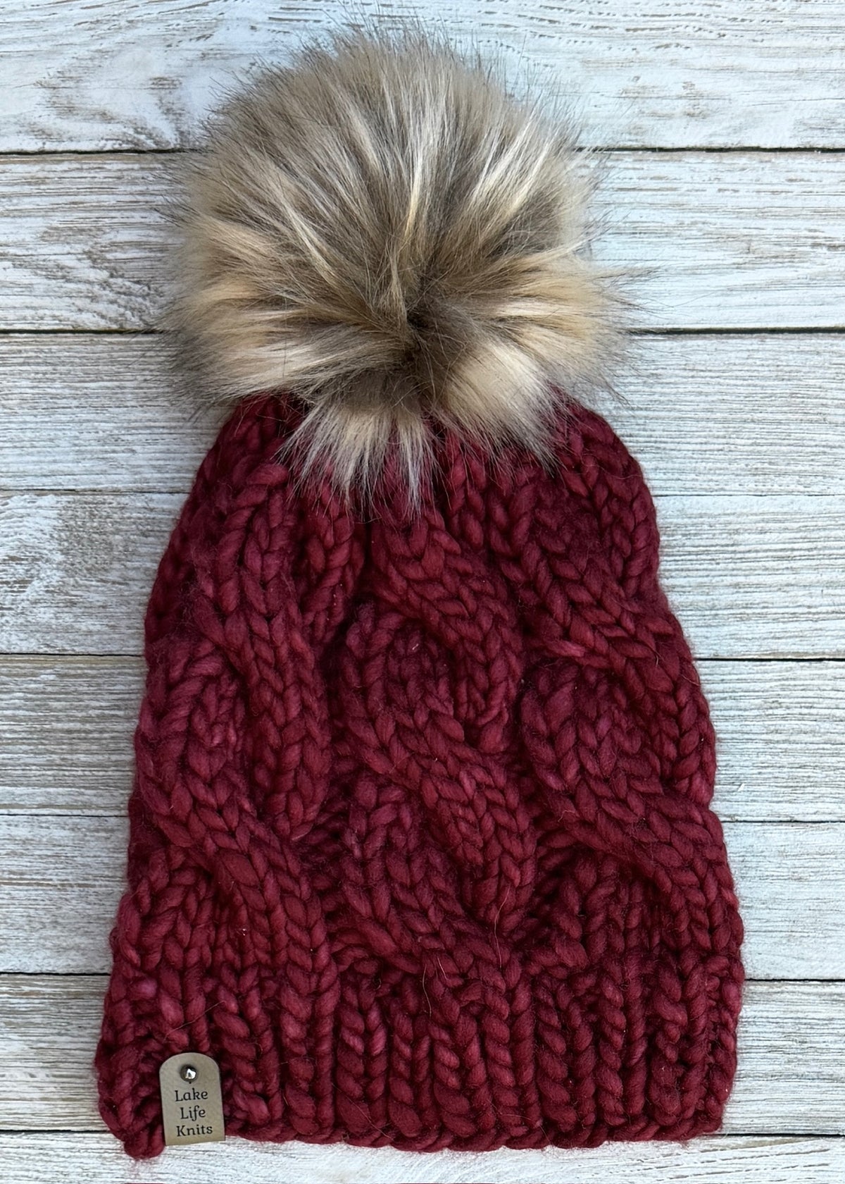 Burgundy hand-knit cable beanie with a brown and cream faux-fur pom and Lake Life Knits tag.